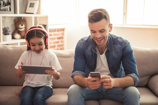 Vater und Tochter sitzen auf der Couch und hören Musik mit Kopfhörern.