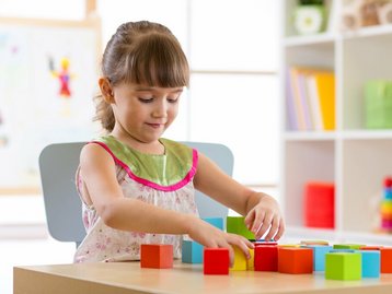 Planung sicher starten mit Hausbau Schutzbrief Mädchen beginnt Bausteine auf Tisch zu stapeln und steht für Planung und Finanzierungssicherheit durch den Hausbau Schutzbrief