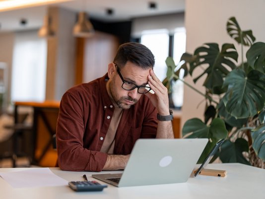 Bauherr verzweifelt wegen Insolvenz beim Hausbau Verzweifelter Bauherr am Tisch mit Laptop, Taschenrechner und Papieren – steht sinnbildlich für finanzielle Belastung durch Bau-Insolvenz ohne passenden Schutz beim Hausbau